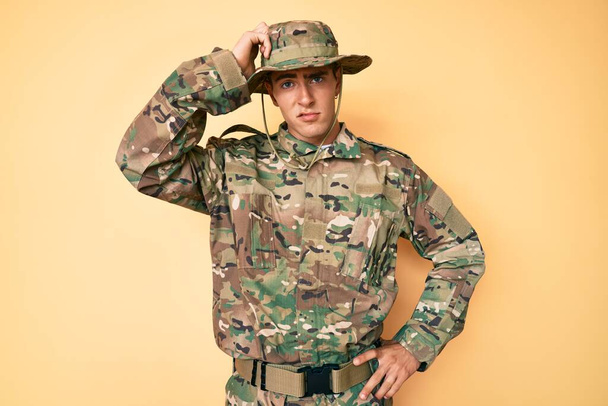 Молодой красивый мужчина в камуфляжной армейской униформе сбивает с толку и задается вопросом. неуверенный в сомнении, думающий с рукой на голове. застенчивое понятие.  - Фото, изображение