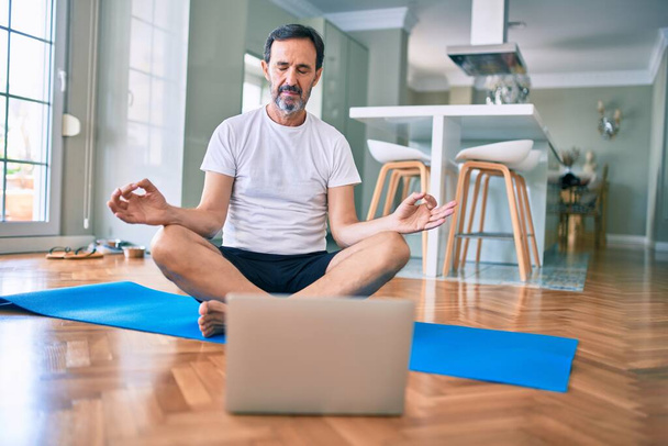 Hombre de mediana edad con entrenamiento de barba y estiramiento haciendo ejercicio en casa mirando videos de yoga en la computadora - Foto, imagen