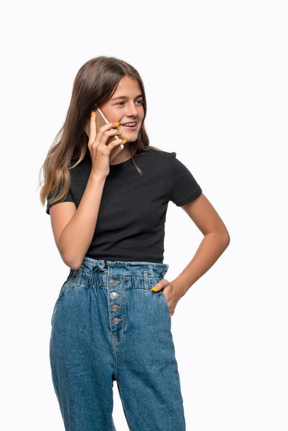 Портрет юной улыбающейся девушки-подростка с брекетами в черной рубашке и джинсах, говорящий по телефону на белом фоне в студии - Фото, изображение