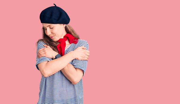 Joven mujer rubia hermosa con boina francesa y camiseta a rayas abrazándose feliz y positivo, sonriendo confiado. amor propio y cuidado personal  - Foto, Imagen