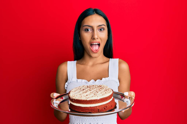Mladá brunetka žena drží mrkvový dort slaví šílený a ohromen pro úspěch s otevřenýma očima křičí vzrušený.  - Fotografie, Obrázek