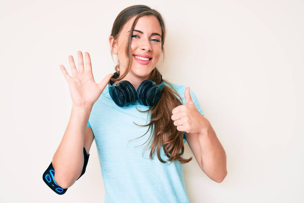 Belle jeune femme caucasienne portant des vêtements de gymnastique et utilisant des écouteurs montrant et pointant vers le haut avec les doigts numéro six tout en souriant confiant et heureux.  - Photo, image