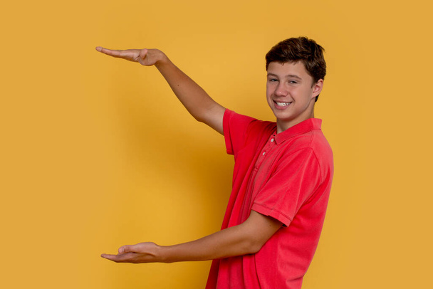 Студийный снимок улыбающегося мальчика, рекламирующего что-то на жёлтом фоне с копировальным пространством - Фото, изображение