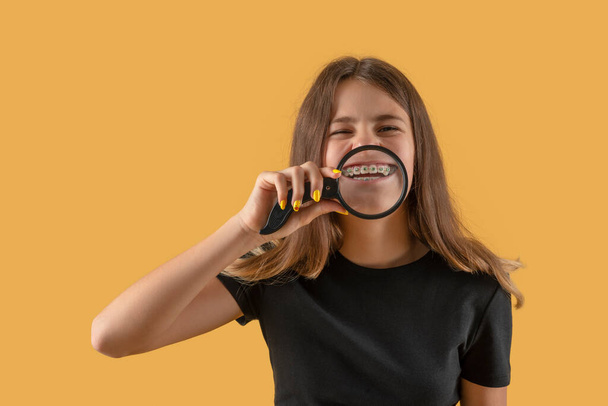 Waist up portrait of an attractive teenager girl with braces  wearing  black shirt playfully demonstrates her teeth through a magnifying glass against yellow background - Foto, Bild