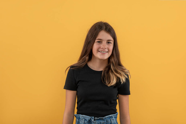 Cintura para cima retrato de uma menina adolescente sorridente atraente com aparelho vestindo camisa preta contra fundo amarelo - Foto, Imagem