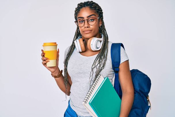 Νεαρή Αφροαμερικανή με κοτσίδες που φοράει φοιτητικό σακίδιο πίνοντας καφέ χαλαρό με σοβαρή έκφραση στο πρόσωπο. απλή και φυσική κοιτάζοντας την κάμερα.  - Φωτογραφία, εικόνα