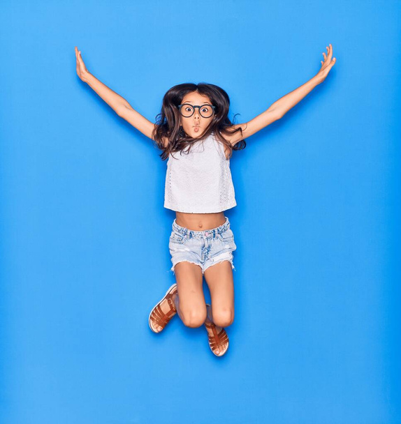 Adorable niña hispana vistiendo ropa casual y gafas sonriendo feliz. Saltando con sonrisa en la cara sobre un fondo azul aislado - Foto, Imagen