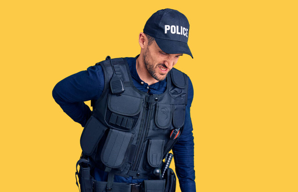 Νεαρός όμορφος άντρας με στολή αστυνομικού που υποφέρει από οσφυαλγία, αγγίζοντας την πλάτη με το χέρι, μυϊκός πόνος  - Φωτογραφία, εικόνα