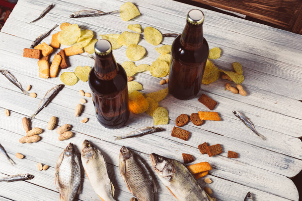 Brown glass bottles of beer and dried fish with chips, nuts, crackers on paper on a white wooden background. Studio photo. - Photo, image