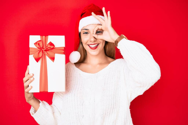 jonge brunette vrouw het dragen van kerst hoed en het houden van een geschenk glimlachen gelukkig doen ok teken met de hand op oog kijken door vingers  - Foto, afbeelding
