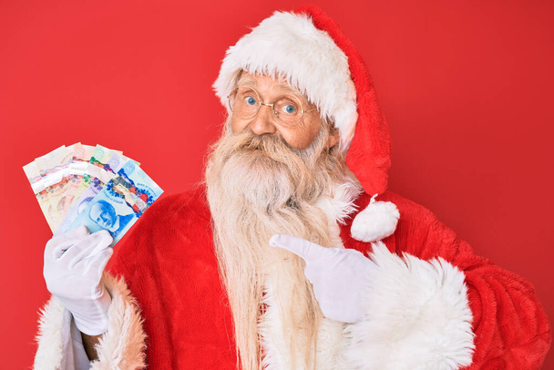 Stary staruszek z siwymi włosami i długą brodą w kostiumie Świętego Mikołaja trzymający kanadyjskie dolary uśmiechnięty szczęśliwy ze wskazaniem ręką i palcem  - Zdjęcie, obraz