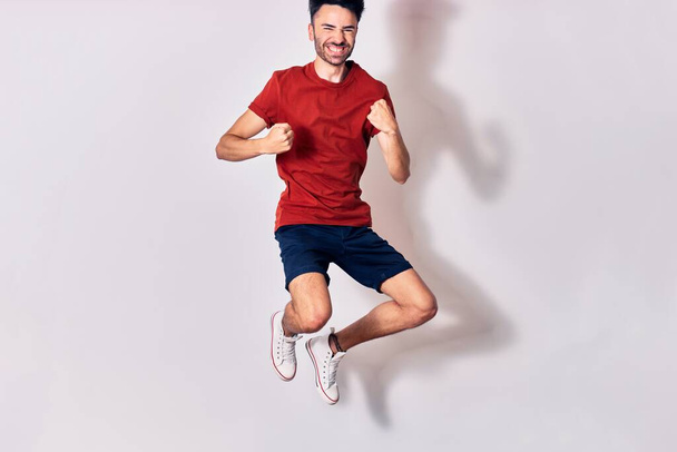 Young handsome man wearing casual clothes smiling happy. Jumping with smile on face doing winner gesture with fists up over isolated white background - Photo, Image