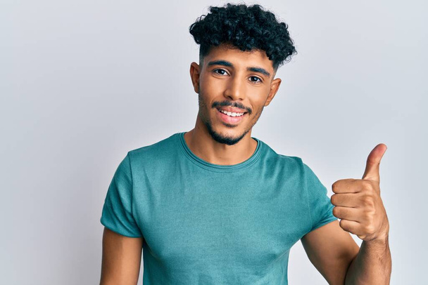 Jonge Arabisch knappe man draagt casual kleding glimlachend gelukkig en positief, duim omhoog doet uitstekend en goedkeuring teken  - Foto, afbeelding
