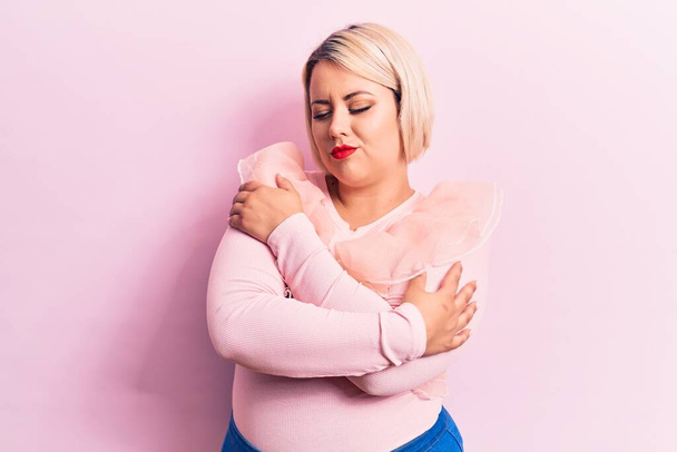 Молодая красивая блондинка плюс размер женщина носит случайный свитер на изолированном розовом фоне обнимая себя счастливым и позитивным, улыбаясь уверенно. Самолюбие и забота о себе - Фото, изображение
