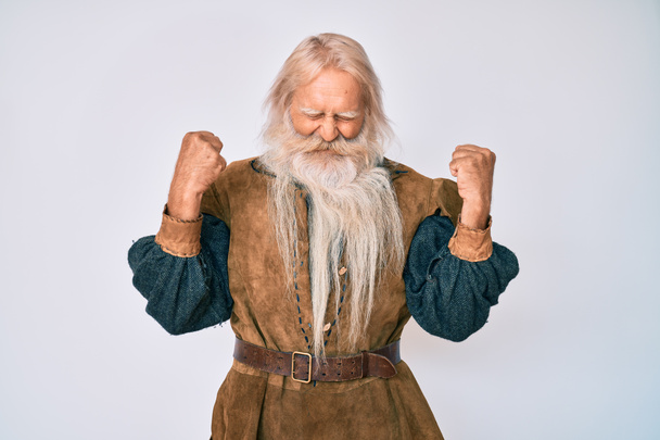 Vieil homme âgé aux cheveux gris et à la barbe longue portant un costume traditionnel viking très heureux et excité faisant geste gagnant avec les bras levés, souriant et criant pour le succès. concept de célébration.  - Photo, image