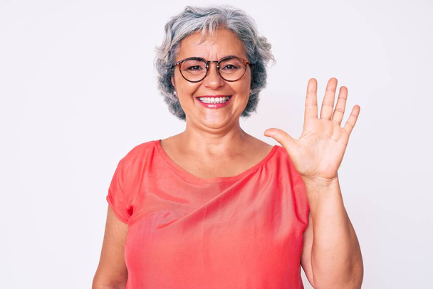 Старшая латиноамериканская серая женщина в повседневной одежде и очках показывает и показывает пальцами номер пять улыбаясь уверенной и счастливой.  - Фото, изображение