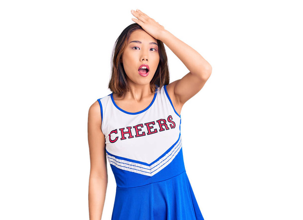 Giovane bella ragazza cinese indossa uniforme cheerleader sorpreso con mano sulla testa per errore, ricordare l'errore. dimenticato, concetto di cattiva memoria.  - Foto, immagini