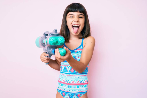 Young little girl with bang wearing swimsuit and watergun sticking tongue out happy with funny expression.  - Photo, Image