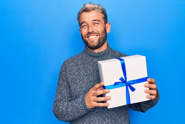 Νέος όμορφος ξανθός άντρας με γενειάδα που κρατάει δώρο γενεθλίων πάνω από απομονωμένο μπλε φόντο που φαίνεται θετικός και χαρούμενος στέκεται και χαμογελά με αυτοπεποίθηση χαμόγελο που δείχνει τα δόντια - Φωτογραφία, εικόνα