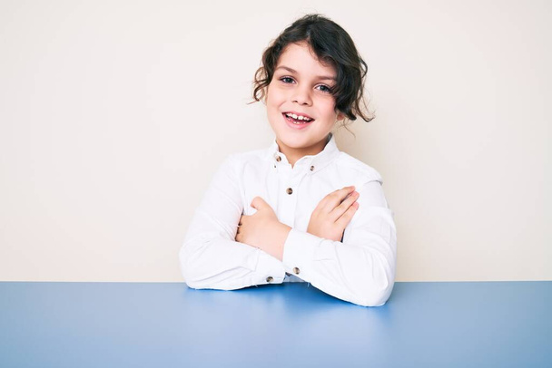 Cute hispanic child wearing casual clothes sitting on the table happy face smiling with crossed arms looking at the camera. positive person.  - Valokuva, kuva