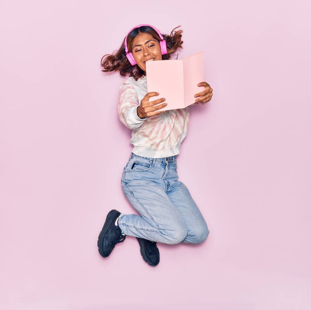 Νεαρή όμορφη Λατίνα γυναίκα ακούγοντας μουσική χρησιμοποιώντας ακουστικά χαμογελώντας ευτυχισμένη. Άλμα με χαμόγελο στο πρόσωπο ανάγνωση βιβλίο πάνω από απομονωμένο ροζ φόντο - Φωτογραφία, εικόνα