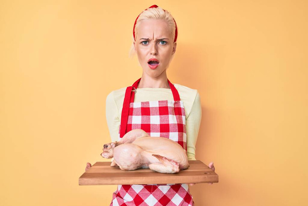 Önlük takan genç sarışın kadın, şok olmuş suratına tavuk pişiriyor, şüpheci ve alaycı görünüyor, ağzı açık kalınca şaşırıyor.  - Fotoğraf, Görsel