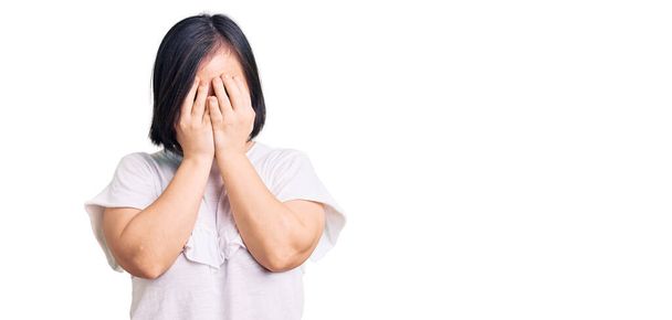 Брюнетка с синдромом Дауна носит повседневную белую футболку с грустным лицом, прикрывающим лицо руками во время плача. депрессия.  - Фото, изображение