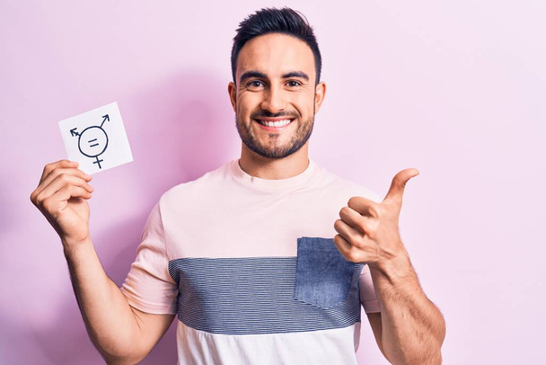 Young handsome man with beard asking for equality holding paper with transgender symbol smiling happy and positive, thumb up doing excellent and approval sign - Photo, Image