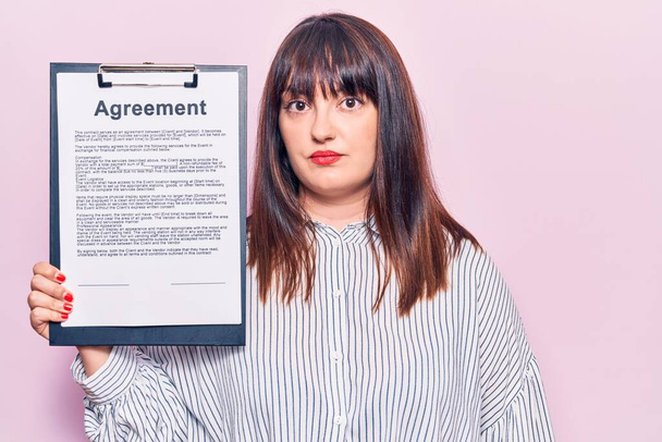 Young plus size woman holding clipboard with agreement document thinking attitude and sober expression looking self confident  - Foto, imagen