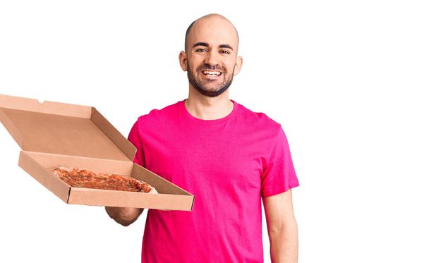 Fiatal jóképű férfi kezében szállítás pizza kartondoboz néz pozitív és boldog állva és mosolyogva magabiztos mosollyal mutatja fogak  - Fotó, kép