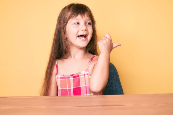 Маленькая кавказская девочка с длинными волосами в повседневной одежде сидит на столе, указывая пальцем вверх в сторону улыбаясь счастливой с открытым ртом  - Фото, изображение
