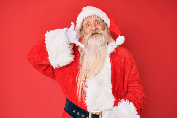Alter älterer Mann mit grauen Haaren und langem Bart in traditionellem Weihnachtsmannkostüm, lächelnd und mit Hand- und Fingernägeln telefonierend. Vermittlung von Konzepten.  - Foto, Bild