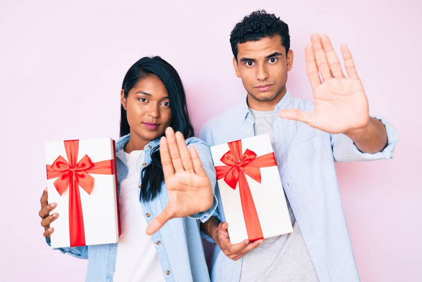 Krásný latinsky mladý pár drží dárek k výročí oslavy s otevřenou rukou dělat stop sign s vážným a sebejistý výraz, obrana gesto  - Fotografie, Obrázek