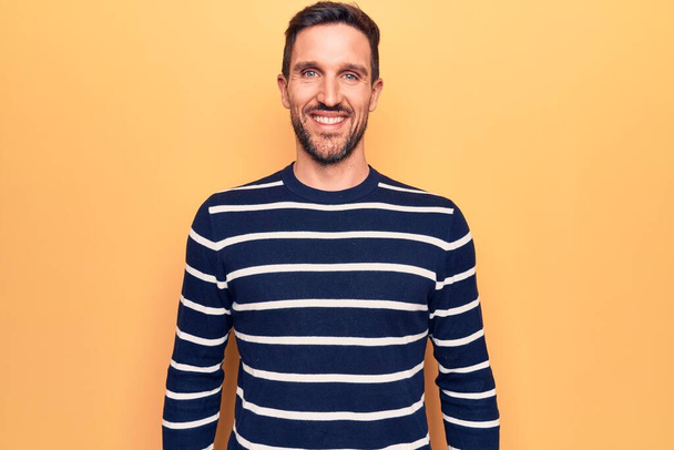 Young handsome man wearing casual striped sweater standing over isolated yellow background with a happy and cool smile on face. Lucky person. - Foto, afbeelding