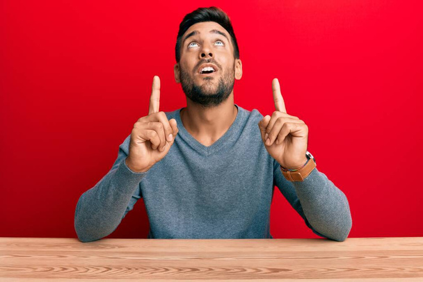 Handsome hispanic man wearing casual style sitting on the table amazed and surprised looking up and pointing with fingers and raised arms.  - Photo, Image