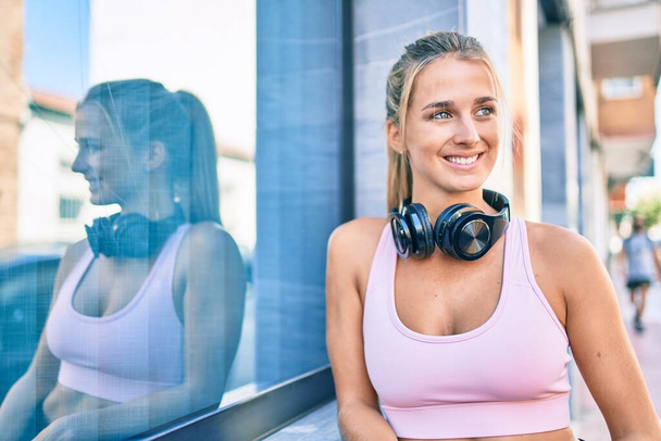 Young blonde sporty girl wearing headphones leaning on the wall at street of city. - Photo, Image