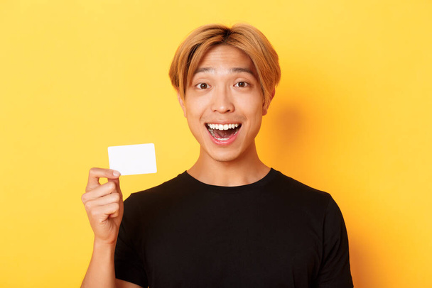 Крупный план взволнованного удивленного симпатичного азиатского парня, улыбающегося изумленного и показывающего кредитку, стоящего на жёлтом фоне - Фото, изображение