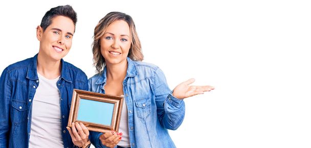 Couple of women holding empty frame smiling cheerful presenting and pointing with palm of hand looking at the camera.  - Foto, Imagen