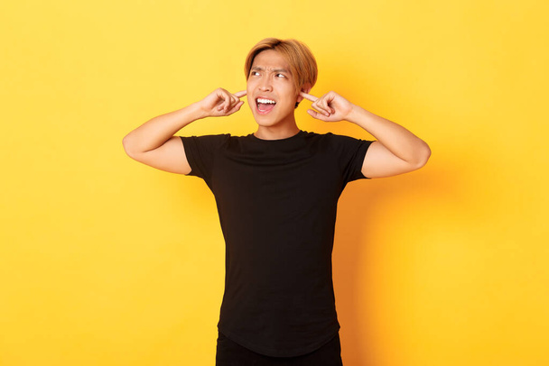 Портрет раздраженного и надоедливого азиатского парня, кричащего, закрытого ушами, потревоженного громким шумом, стоящего на желтом фоне - Фото, изображение