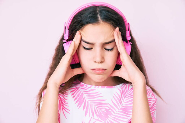 Cute hispanic child girl listening to music using headphones with hand on head, headache because stress. suffering migraine.  - Photo, Image
