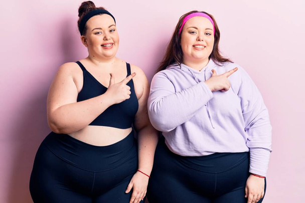 Young plus size twins wearing sportswear cheerful with a smile of face pointing with hand and finger up to the side with happy and natural expression on face  - Photo, Image