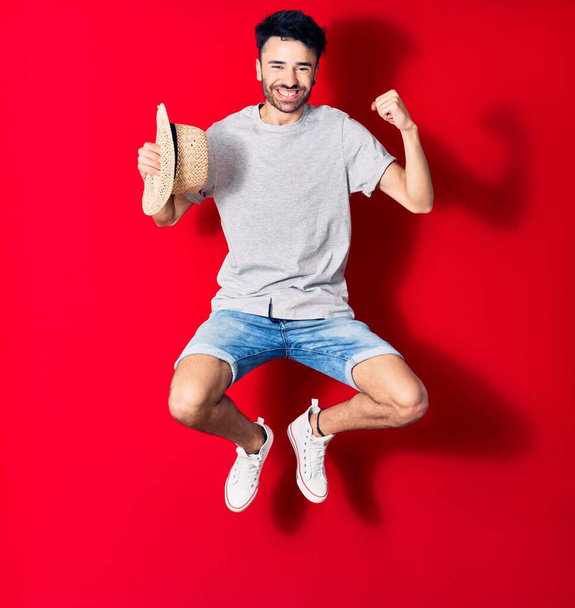 若いハンサムなヒスパニック系の男性が幸せな笑顔カジュアルな服を着ている。顔に笑顔でジャンプ開催夏の帽子お祝いで拳で上の孤立した赤の背景. - 写真・画像