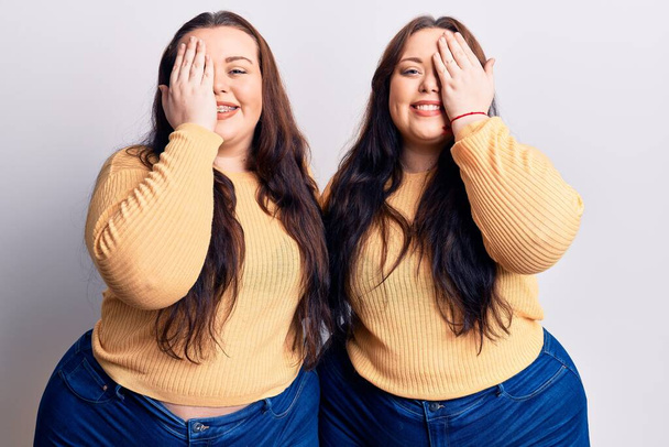 Young plus size twins wearing casual clothes covering one eye with hand, confident smile on face and surprise emotion.  - Fotografie, Obrázek