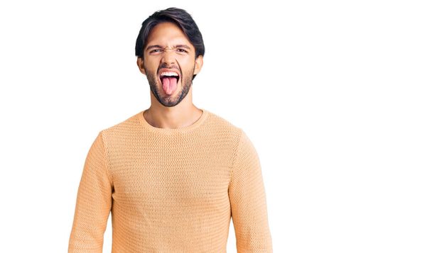 Een knappe Spaanse man met een nonchalante trui die zijn tong uitsteekt, blij met een grappige uitdrukking. emotie concept.  - Foto, afbeelding