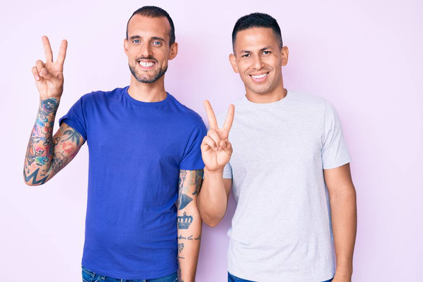 Nuori homo pari kaksi miestä päällään rento vaatteita hymyillen onnellinen kasvot silmää kameran tekee voitonmerkki. Numero kaksi..  - Valokuva, kuva