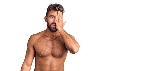Fiatal spanyol férfi fürdőruhát visel félmeztelenül, egyik szemét kézzel takarja, magabiztos mosollyal az arcán és meglepetésszerű érzelmekkel..  - Fotó, kép