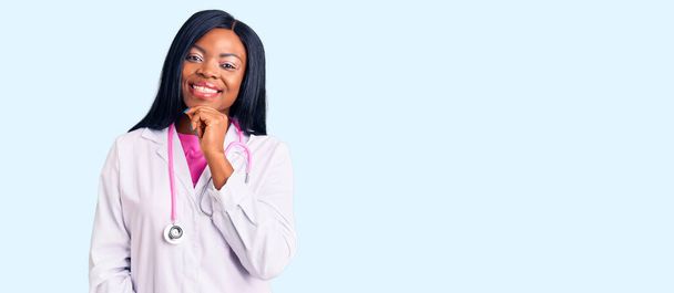 Młoda Afroamerykanka w stetoskopie wyglądająca pewnie przed kamerą, uśmiechnięta ze skrzyżowanymi ramionami i ręką podniesioną na brodzie. pozytywne myślenie.  - Zdjęcie, obraz