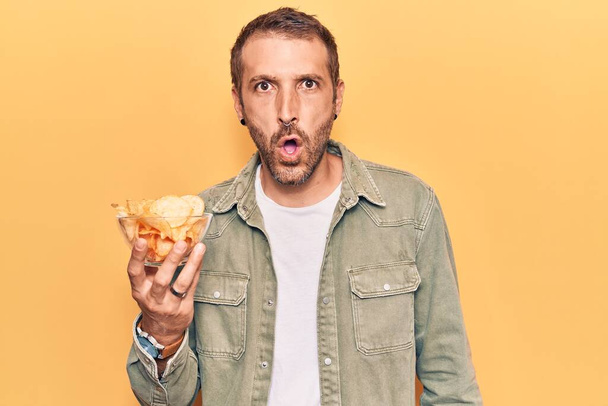 Молодой красивый мужчина держит картофельный чип испуганный и изумленный с открытым ртом для удивления, лица неверия  - Фото, изображение