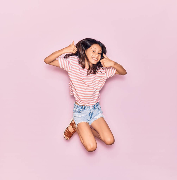Αξιολάτρευτο ισπανόφωνο κορίτσι που φοράει καθημερινά ρούχα χαμογελώντας χαρούμενο. Άλμα με χαμόγελο στο πρόσωπο κάνει εντάξει σημάδι με τους αντίχειρες πάνω από απομονωμένο ροζ φόντο - Φωτογραφία, εικόνα