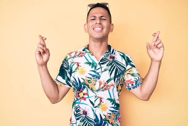 Homem latino bonito jovem vestindo roupas de verão gesto dedo cruzado sorrindo com esperança e olhos fechados. sorte e conceito supersticioso.  - Foto, Imagem
